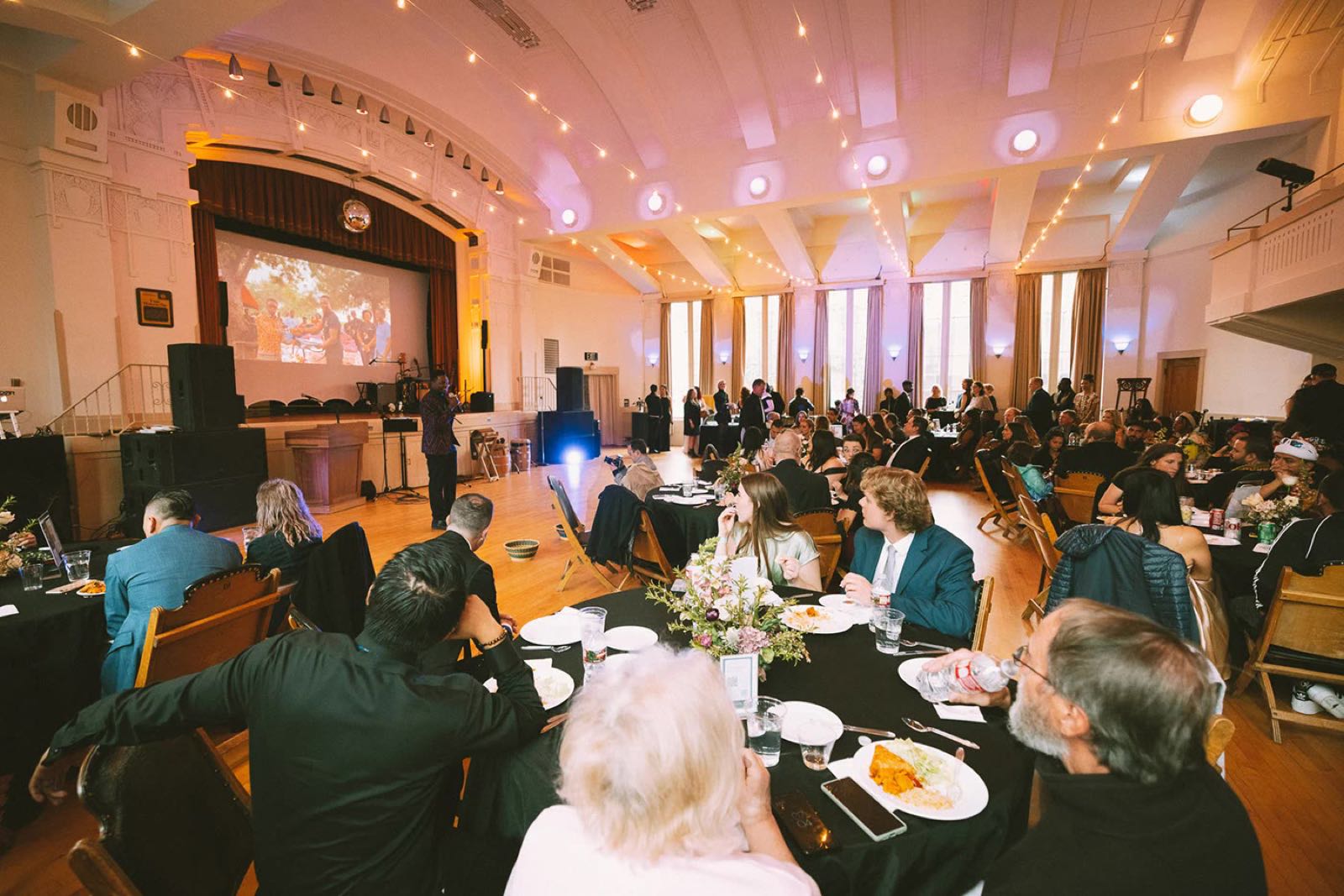 Guests at round tables in a grand hall as Godwill Tandoh speaks on stage at a Dreamers Wish gala